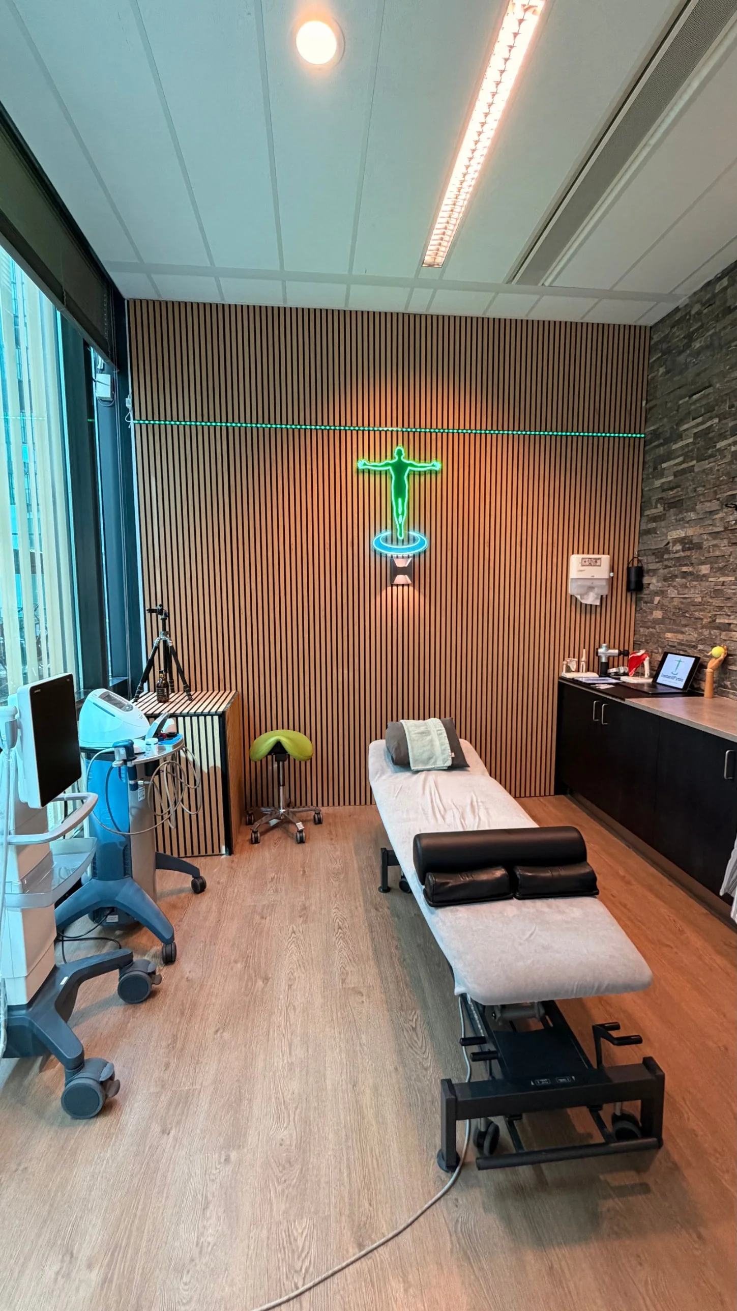 Behandelkamer van InstantFysio met echografie- en shockwaveapparatuur en het verlichte logo op een eiken lattenwand